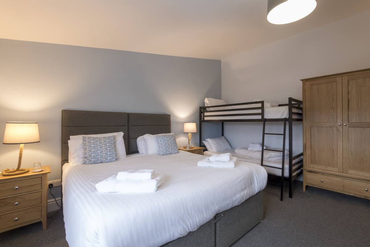 Una cama bien hecha con sábanas y almohadas blancas, una lámpara de noche, una cama litera y un armario de madera.