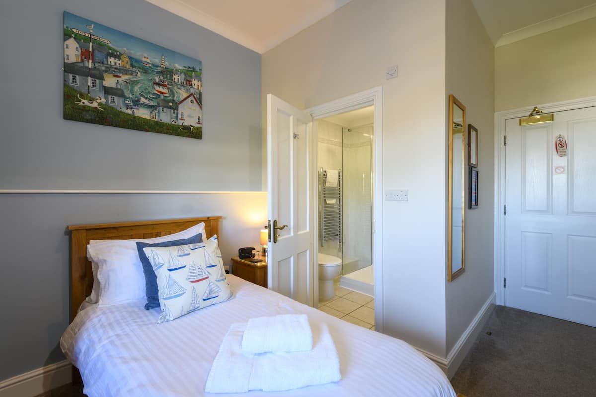 Una cama bien hecha con sábanas blancas y un cojín con diseños de veleros, un cuadro de una escena costera en la pared, y un baño con puerta de cristal y un inodoro blanco.