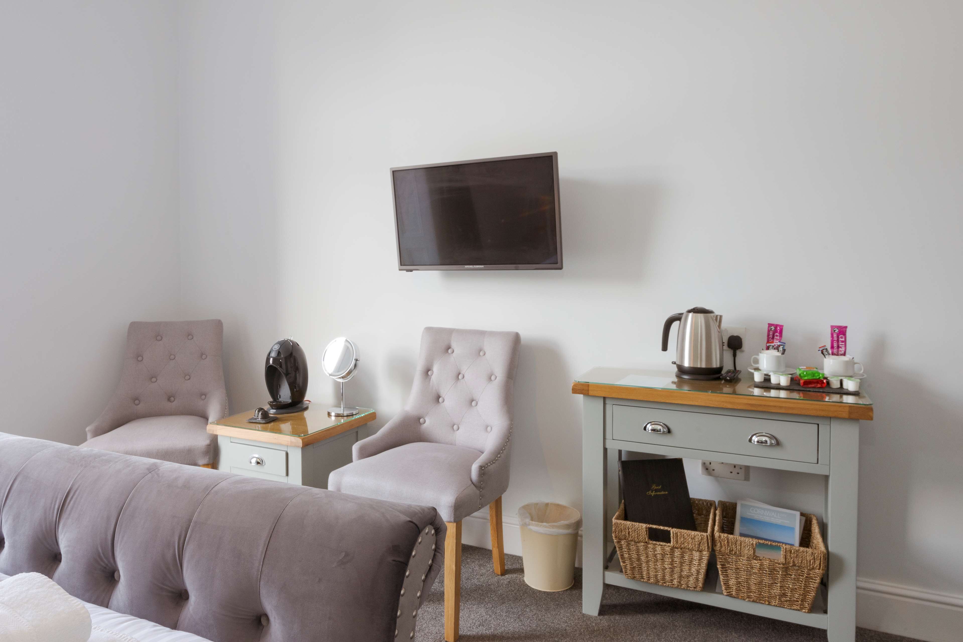 A neatly arranged hotel room with a bed, two armchairs, a small table with a kettle, cups, and a mirror. There is a TV mounted on the wall and a wooden cabinet with baskets.