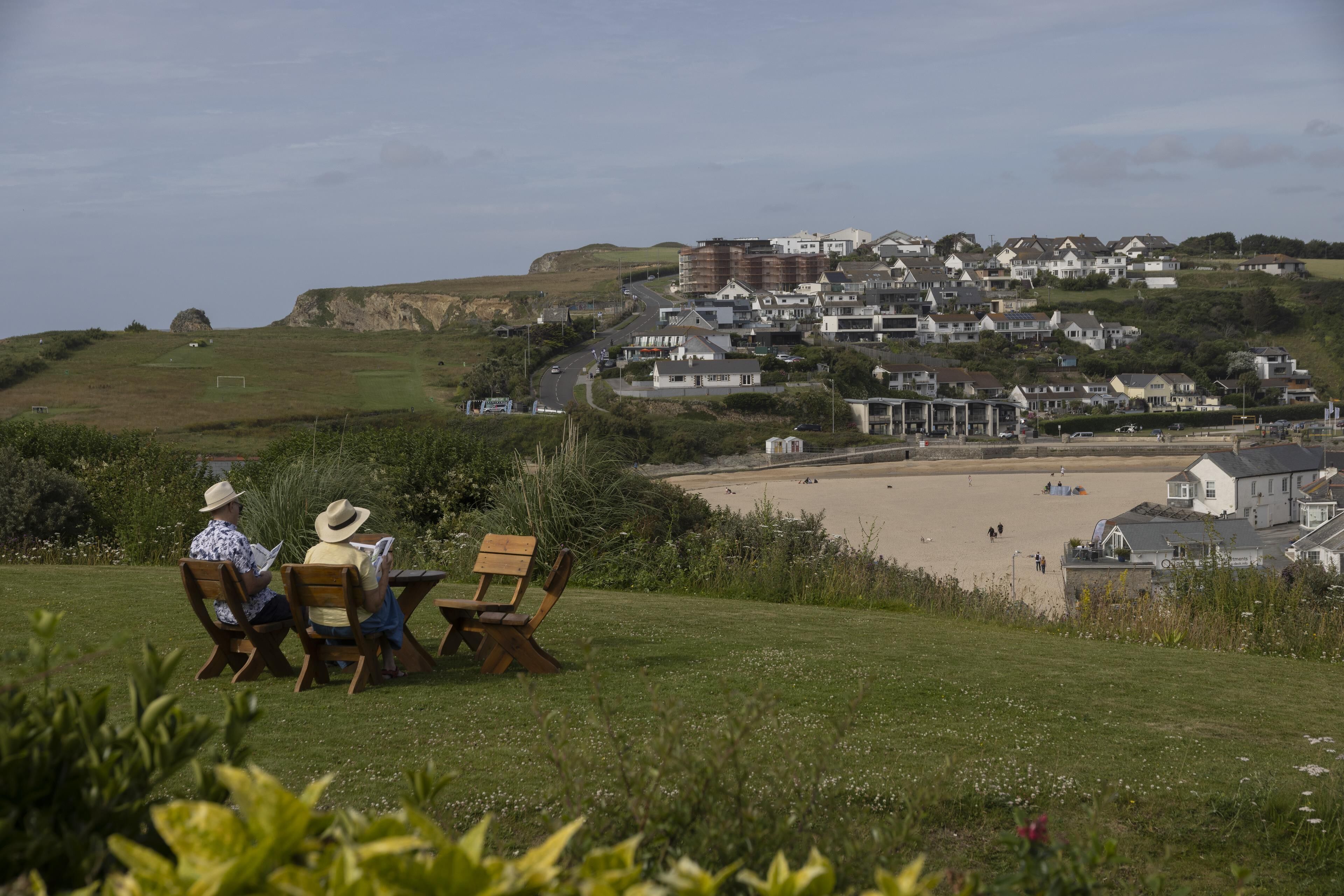 A coastal residential area with houses, a beach, and a grassy hill with people sitting on benches.