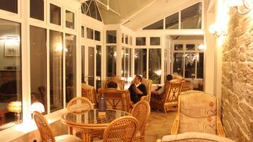 A covered outdoor seating area with wicker chairs and tables, large windows, and wall-mounted lights.