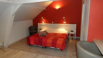 Een slaapkamer met een schuine plafond, een bed met een rode en oranje spreideken, twee bedlampjes, een stoel en een klein tafeltje.