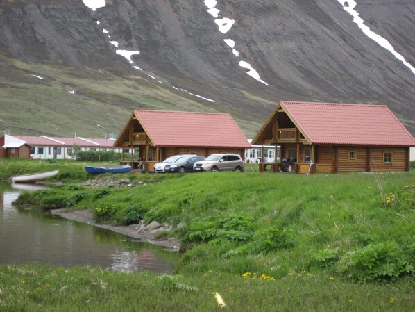 A row of wooden cabins with red roofs, a small stream, and a mountainous backdrop.