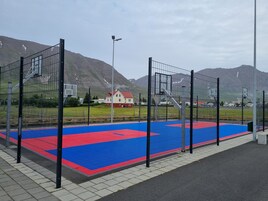 An outdoor basketball court with a red and blue playing surface, surrounded by a metal fence.