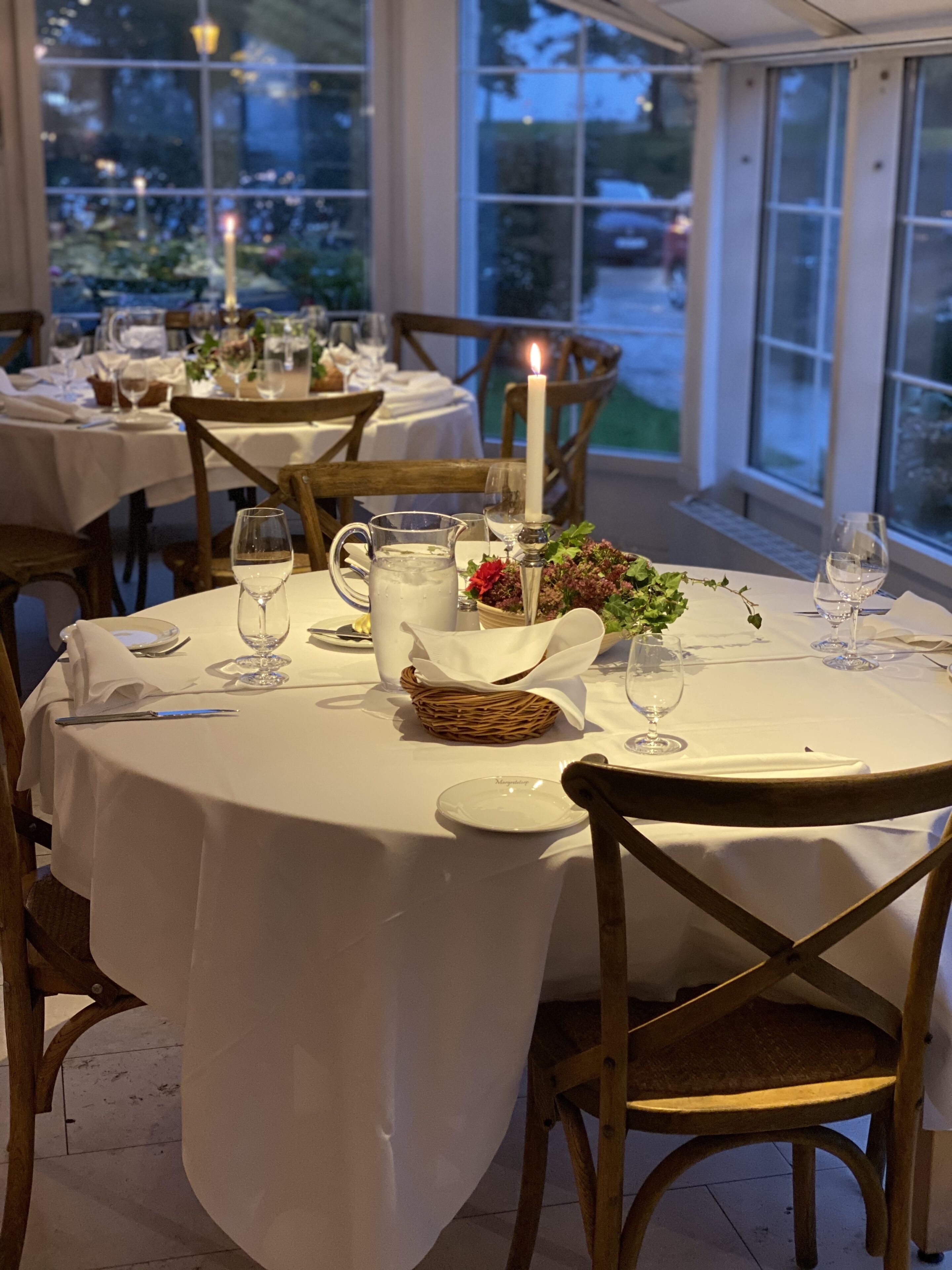 Un comedor con mesas redondas dispuestas para una comida, con manteles blancos, sillas de madera y un centro de mesa con velas.