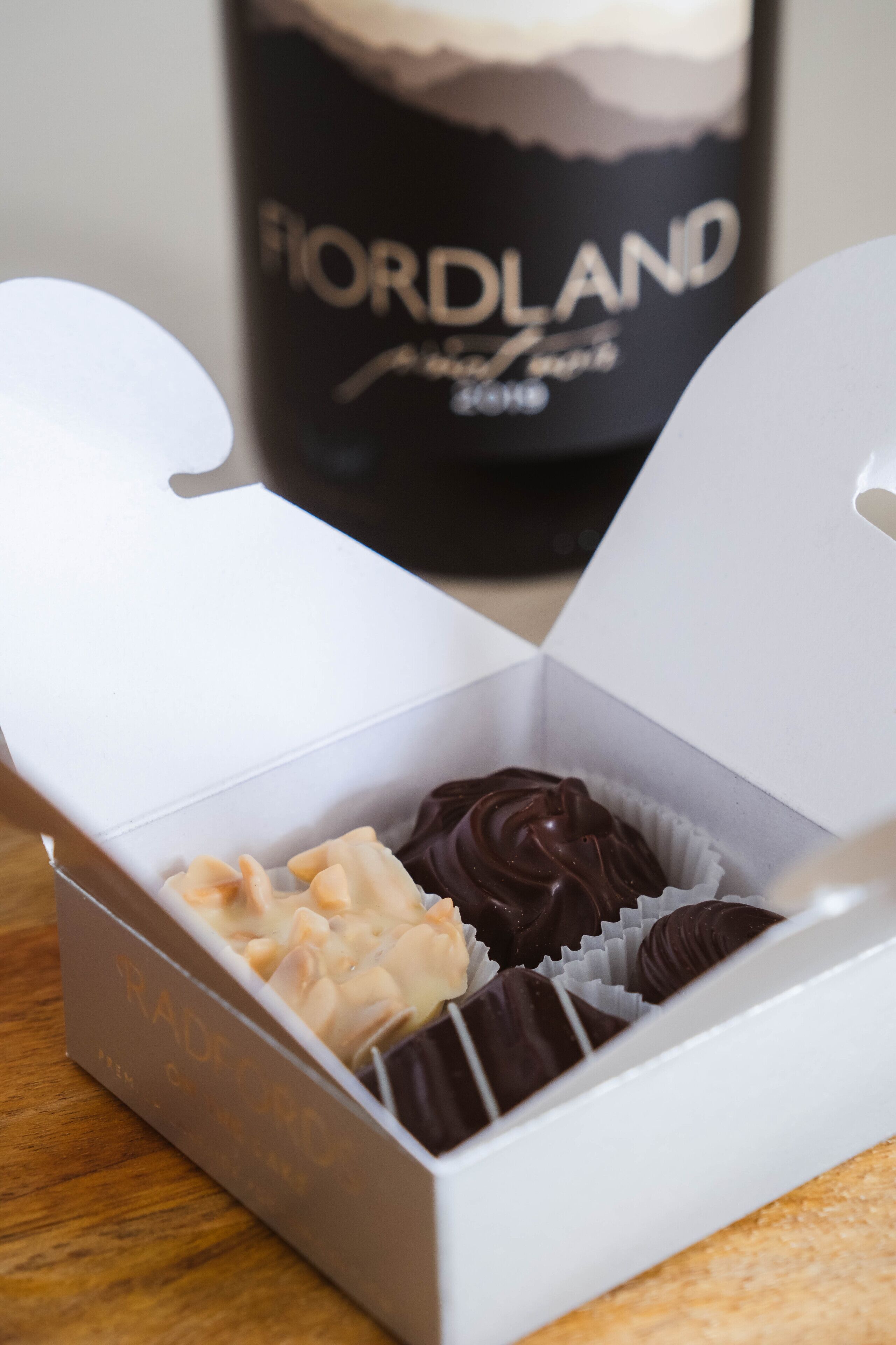 Une boîte blanche contenant trois chocolats, avec une bouteille portant l’étiquette « FIORDLAND » en arrière-plan.