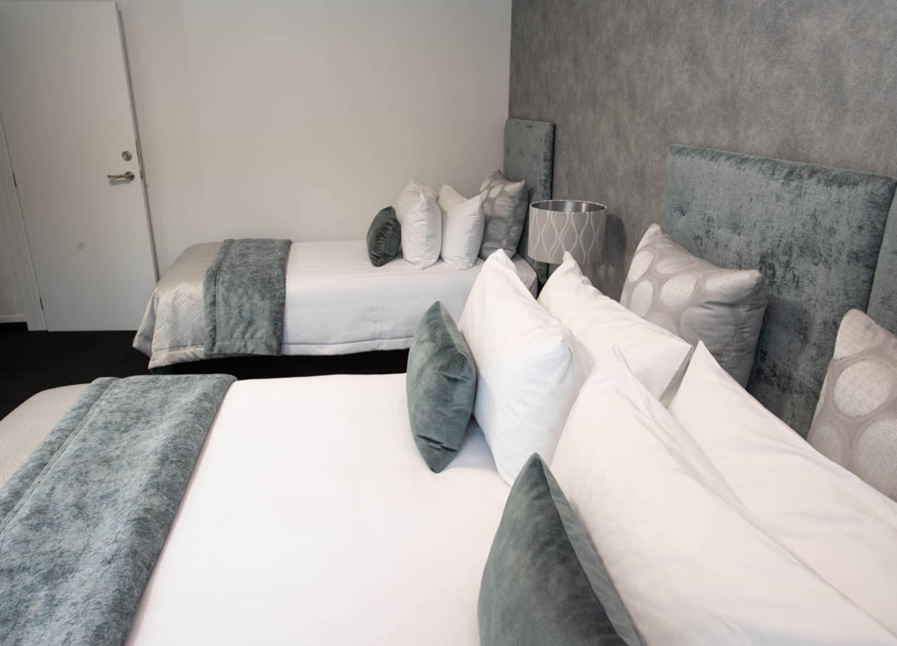 A hotel room with two beds, each with a grey blanket and pillows, a bedside table with a lamp, and a grey headboard.