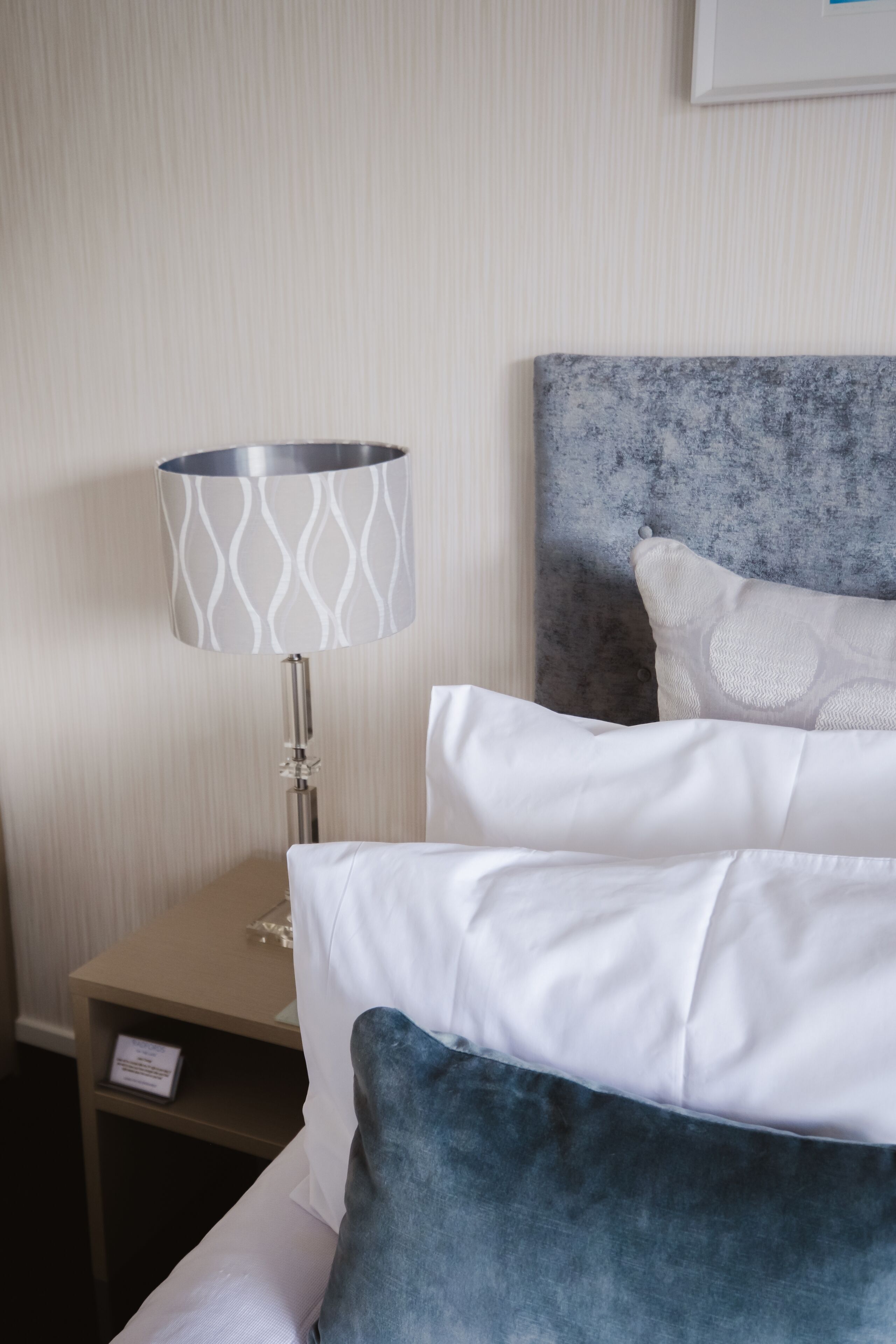 A neatly made bed with white and blue pillows, a metal-framed lamp with a patterned shade, and a small bedside table with a sign.