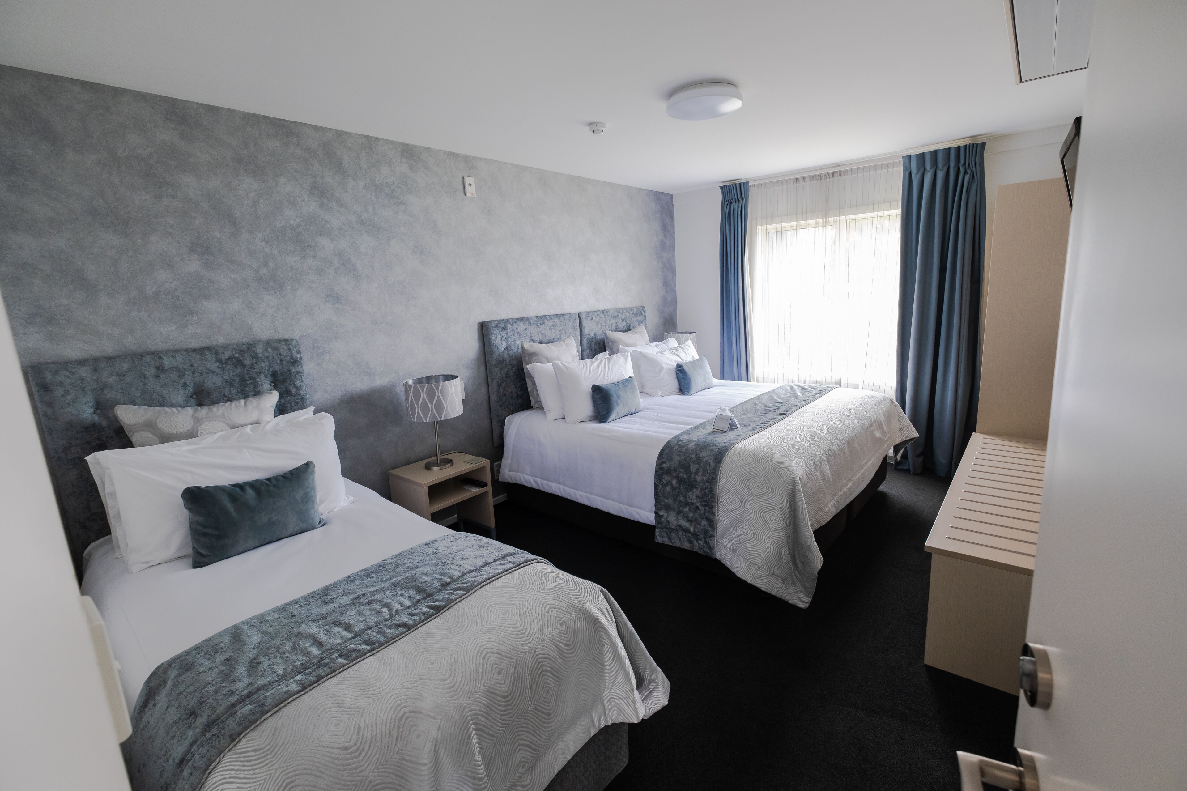 A hotel room with two beds, a bedside table with a lamp, a wardrobe, and a window with curtains.