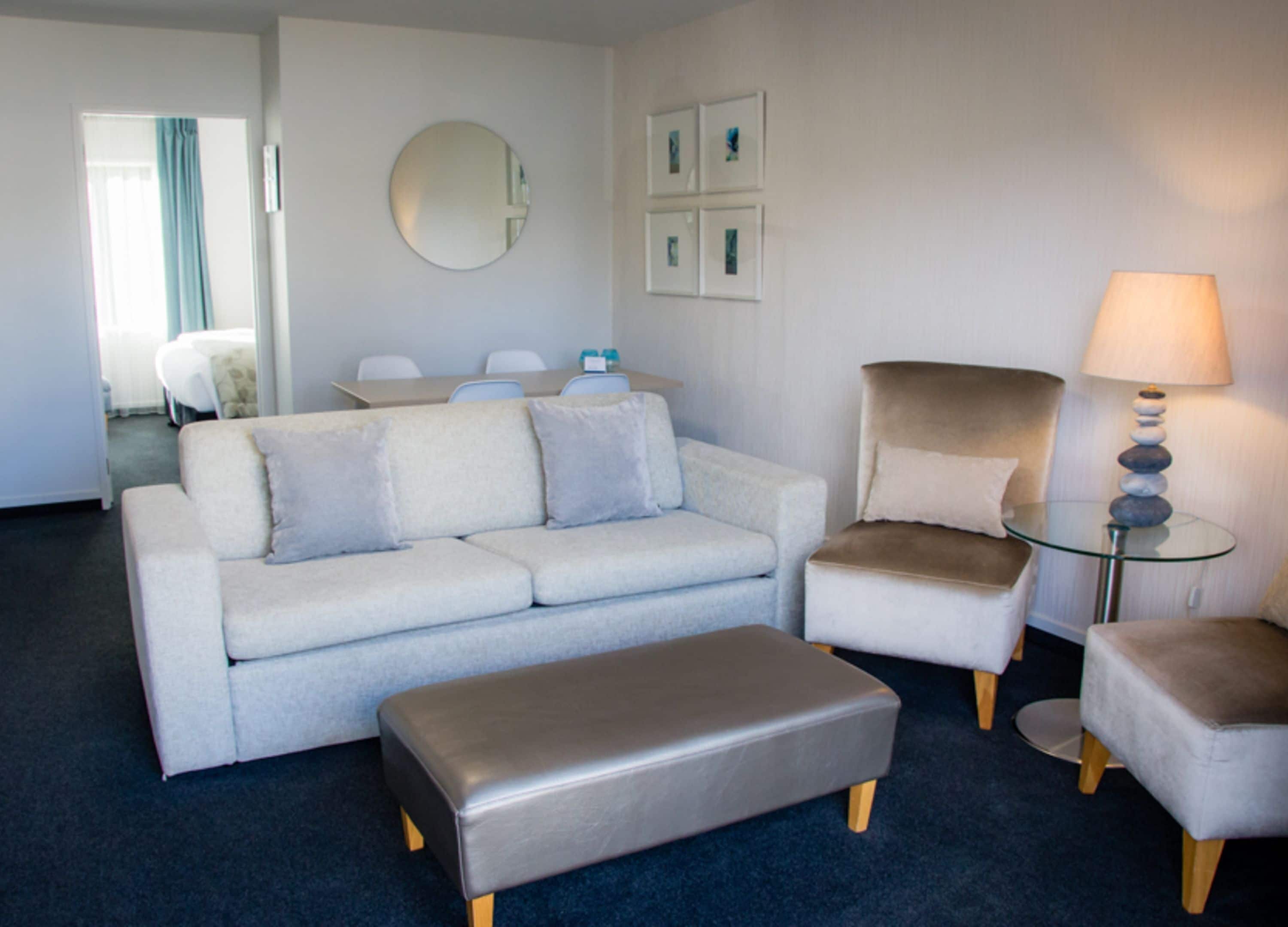 Un salon moderne avec un canapé gris, un fauteuil assorti et une table basse en verre. Un miroir rond est accroché au mur, et des œuvres d’art encadrées se trouvent au-dessus.