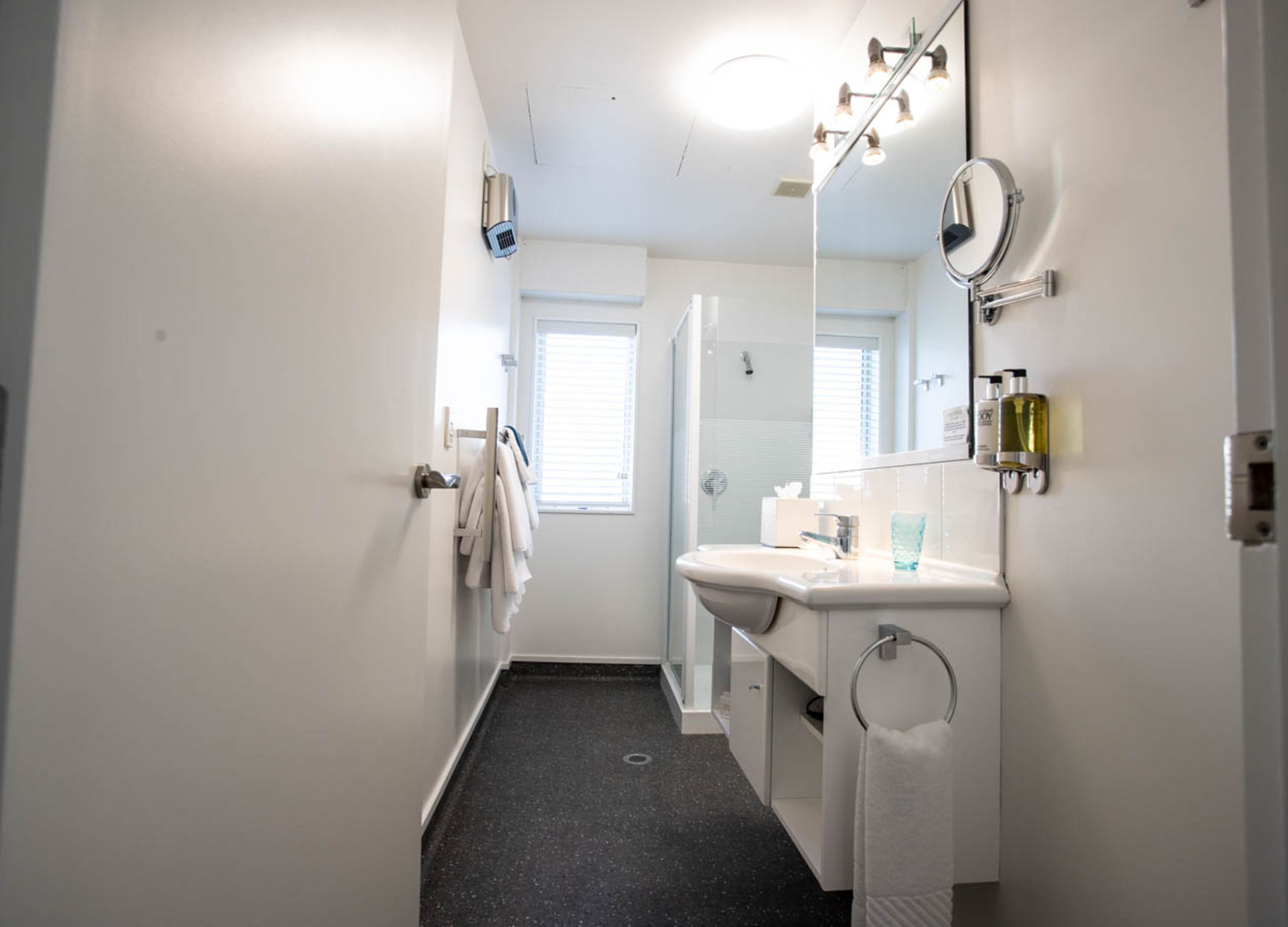 Een smalle, goed verlichte gang die leidt naar een badkamer met een witte wastafel, spiegel en handdoekhouder.