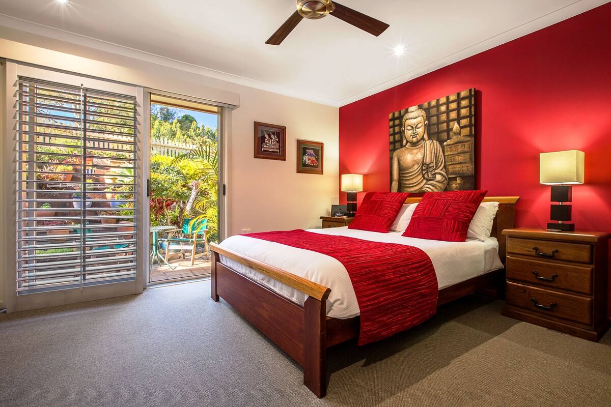 Un dormitorio con una cama grande, ventilador de techo, pared de acento roja y vista a un jardín a través de una puerta corrediza.