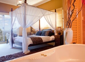 A bedroom with a four-poster canopy bed, a large window with a view, a bedside table with a lamp, and a large white vase. - Avocado Sunset B&B (Tamborine Mountain)