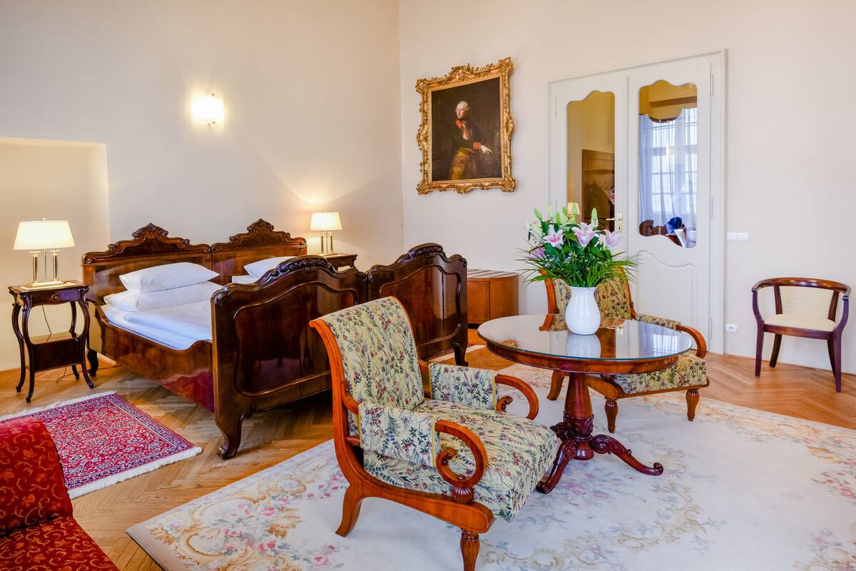 Een kamer met een bed, twee stoelen, een tafel met een vaas en een spiegel.