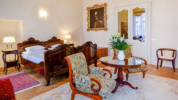 A room with a bed, two chairs, a table with a vase, and a mirror.