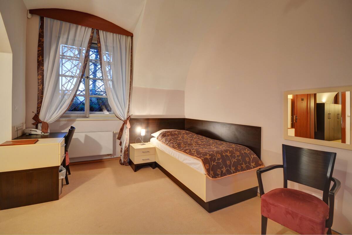 Een slaapkamer met een bed, een bureau, een stoel, een nachtkastje en een raam met gordijnen.