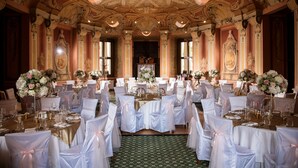 Une salle de banquet ornée de moulures au plafond, de lustres et de compositions florales. Les tables sont dressées avec des nappes blanches et des chaises.