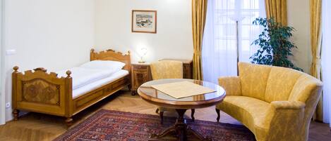 Una habitación con una cama de madera, una mesa redonda, un sillón amarillo, una mesita con una lámpara, un cuadro en la pared y un ventanal con cortinas.