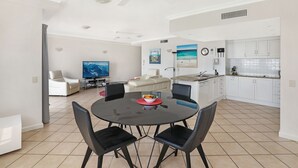 Une cuisine moderne avec des meubles blancs, un coin repas doté d’une table ronde et de chaises noires, et un salon avec un téléviseur et des fauteuils.