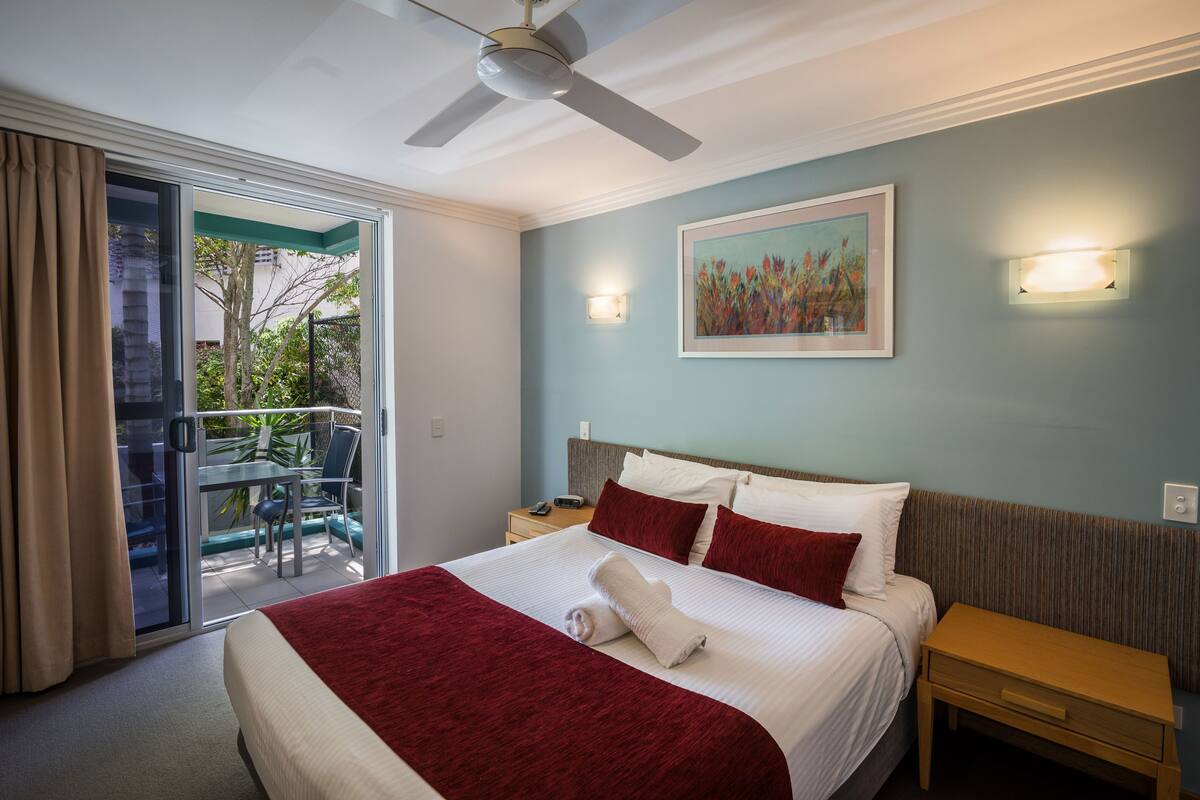A hotel room with a bed, a nightstand, a ceiling fan, a painting on the wall, and a balcony with a table and chairs.