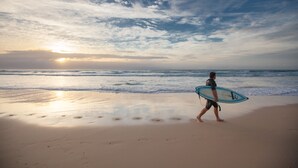 Une personne marchant sur une plage, une planche de surf à la main, avec les vagues de l’océan et un coucher de soleil en arrière-plan.