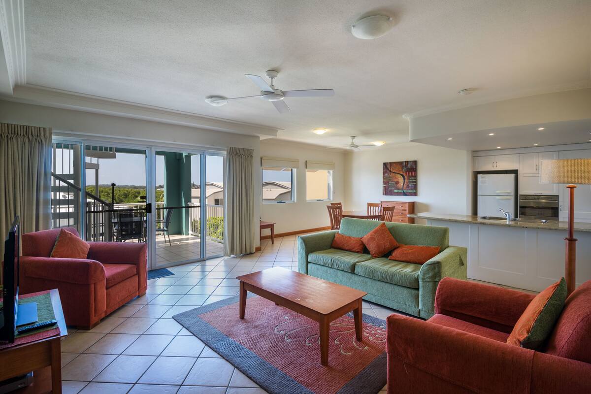 A spacious living area with a red sofa, a green sofa, and a wooden coffee table. There is a balcony with a view of the outdoors.