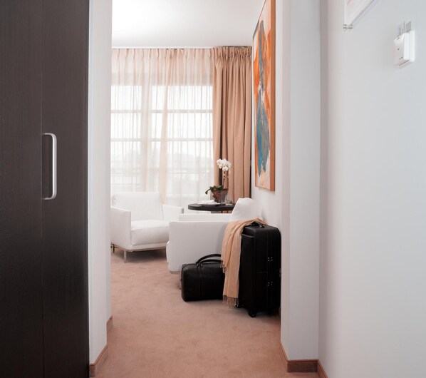 A hotel room with a dark wooden door, a white sofa, a small table with a vase of flowers, a black suitcase, and a beige carpet.