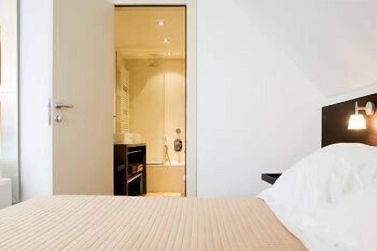 A hotel room with a bed, a nightstand, a shelf with toiletries, and a door leading to a bathroom.