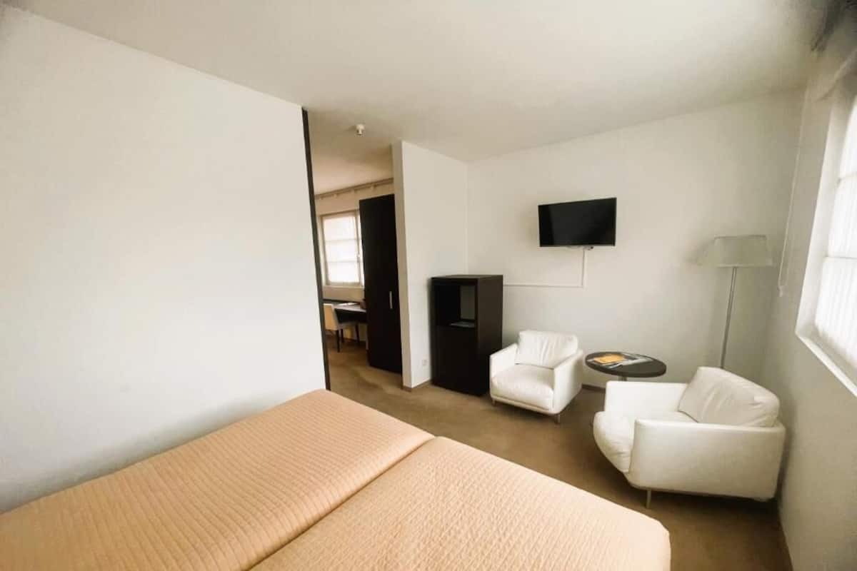 A hotel room with a bed, two white armchairs, a desk, a TV, and a lamp.