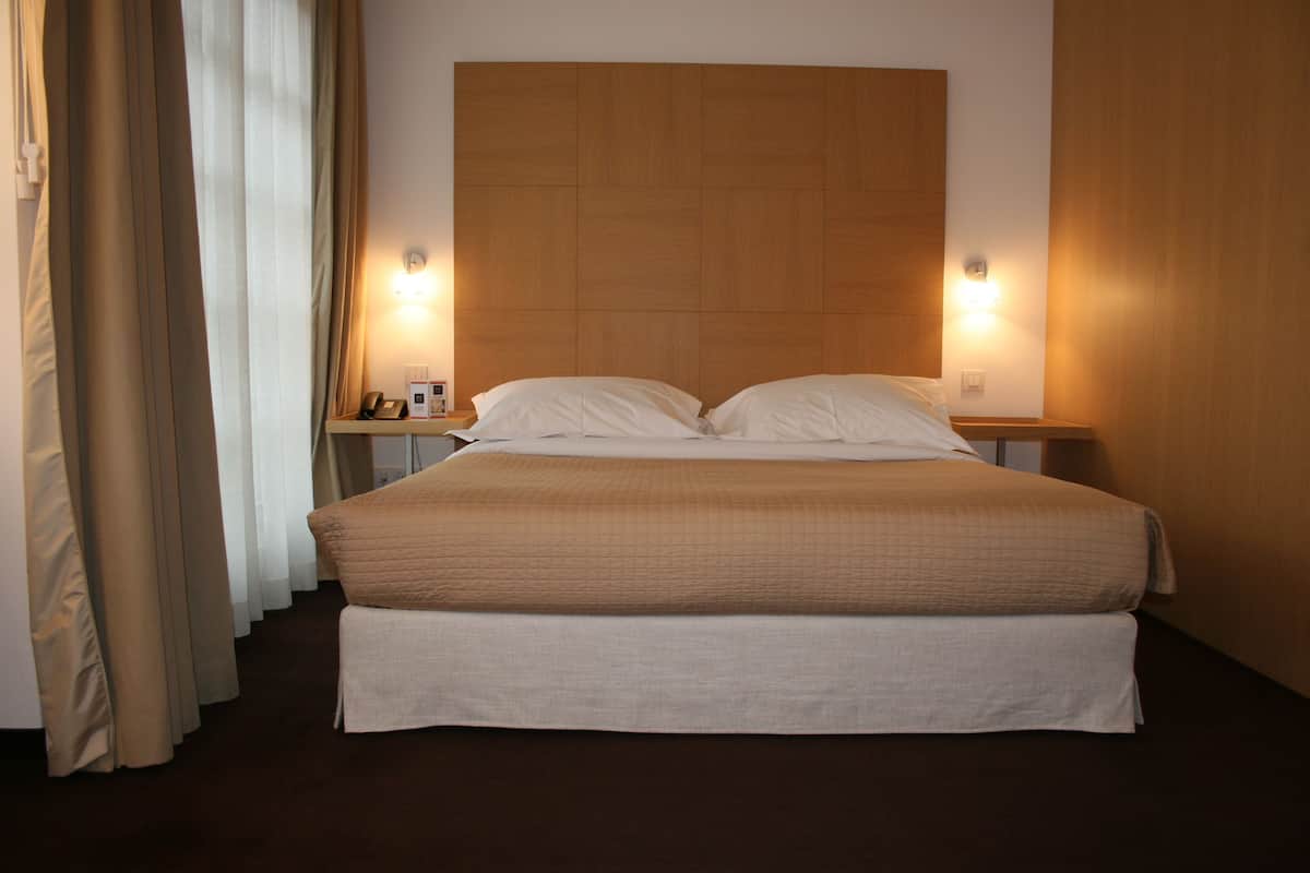 A hotel room with a large bed, two bedside tables, a nightstand, and a curtain.