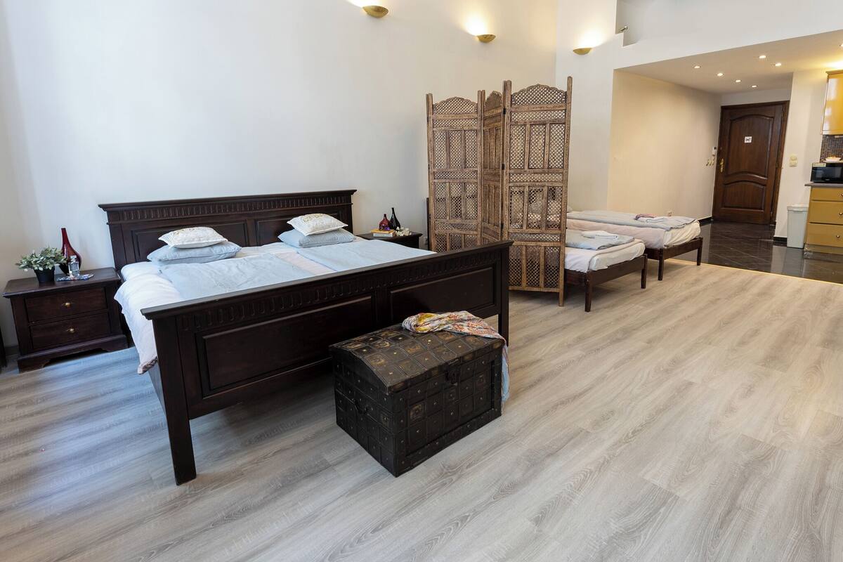 A bedroom with a large bed, two bedside tables, a chest of drawers, a wardrobe, and a small kitchenette.