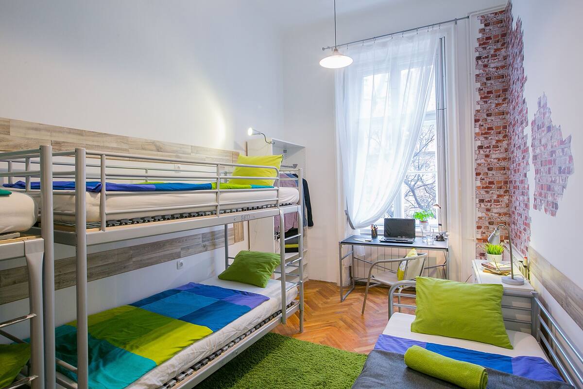 A dormitory room with bunk beds, a desk, a chair, a laptop, and a window with curtains.