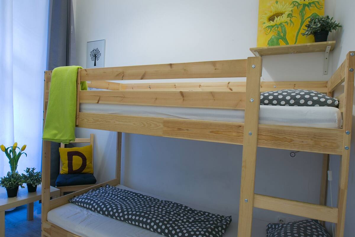 A bunk bed with a shelf above it, a small table with a plant, and a window with curtains.