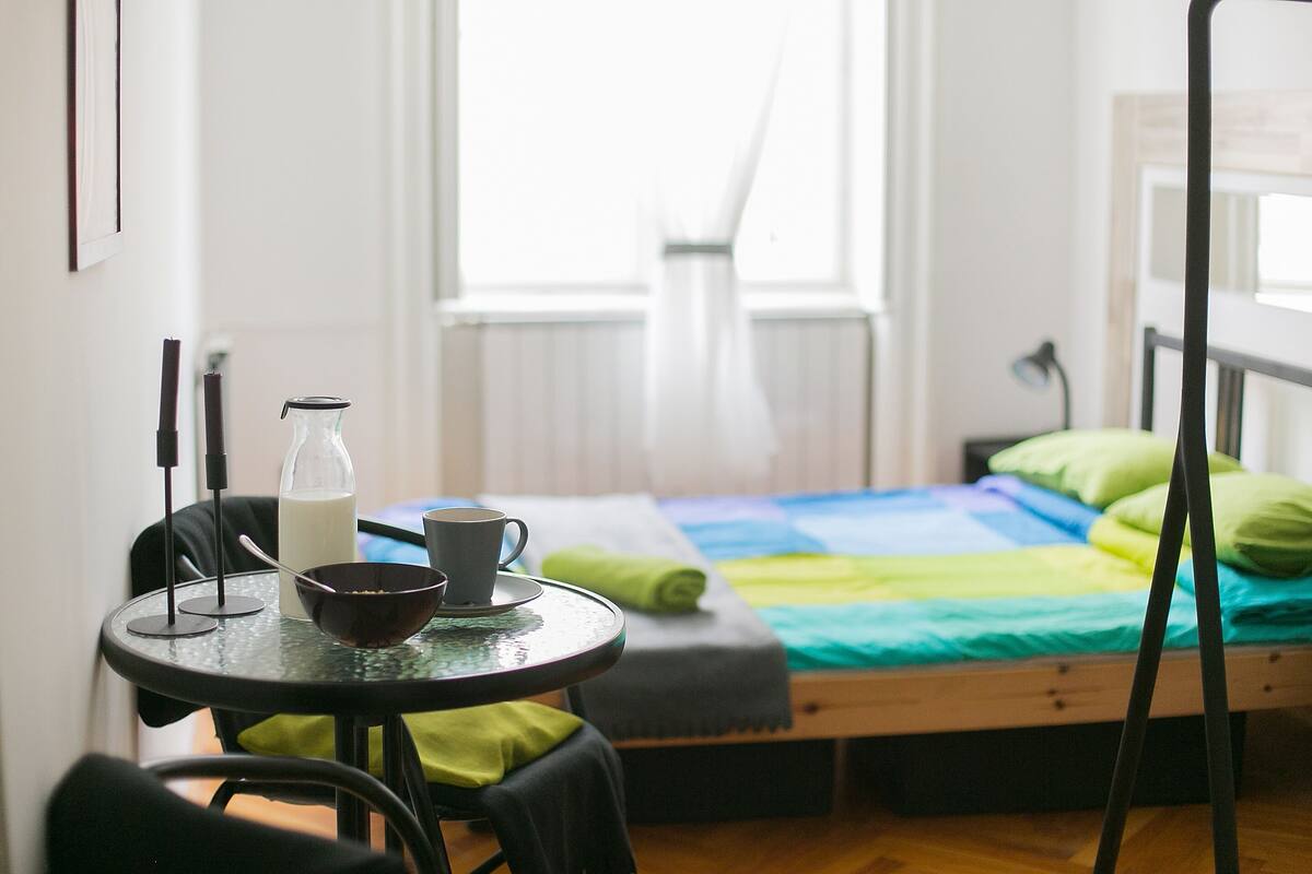 A small room with a bed, a table with a cup and a bottle, and a chair.