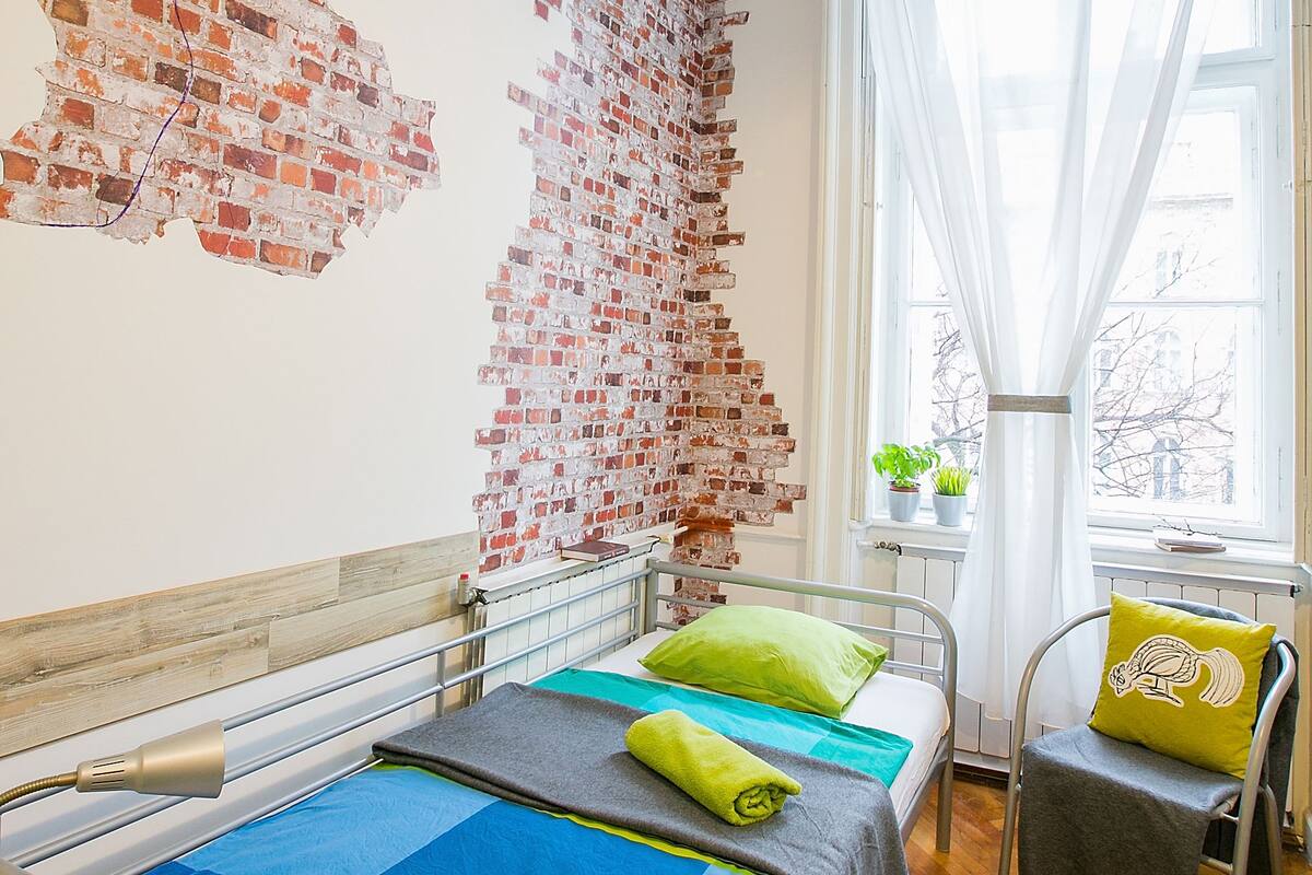A room with a bed, a chair, a window with sheer curtains, and a brick wall mural.
