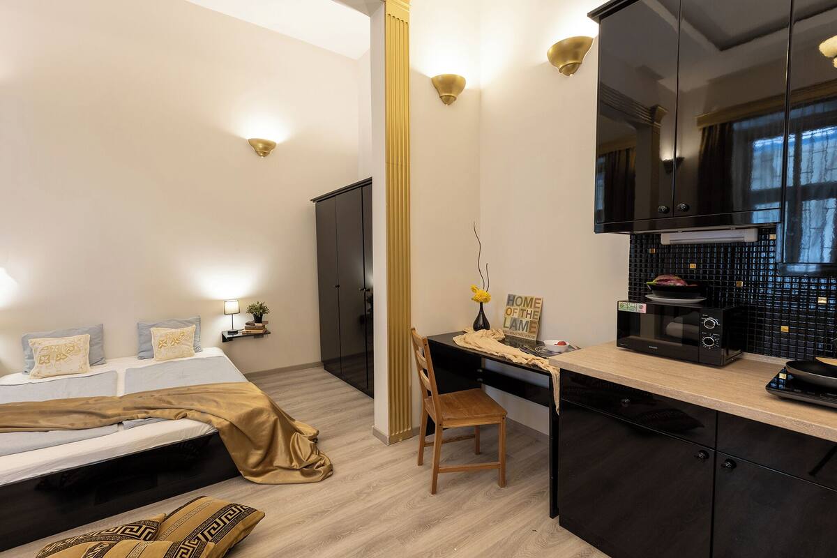 A compact bedroom with a bed, bedside table, and a small desk with a chair. A kitchen area with a microwave and cabinets is visible.