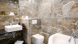 Et moderne badeværelse med marmorbeklædt væg, en hvid vask, toilet og et fristående badekar.