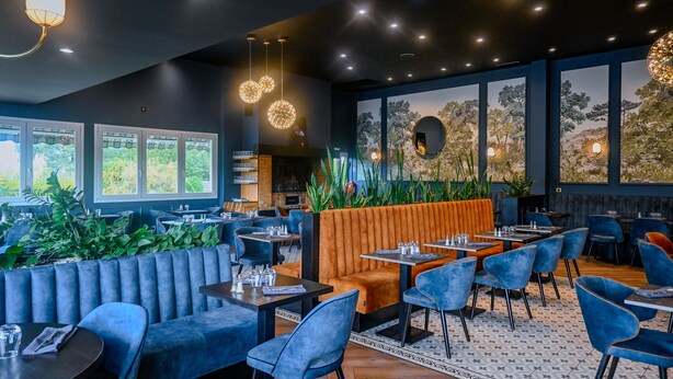 En moderne restaurant med blå og orange siddepladser, store vinduer og et dekorativt loft.
