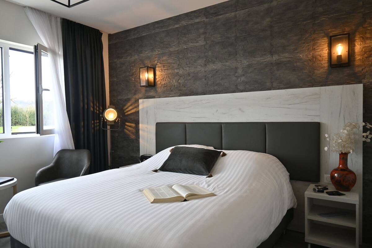 A modern hotel room with a large bed, a bedside table with a vase, and a view of the outdoors.