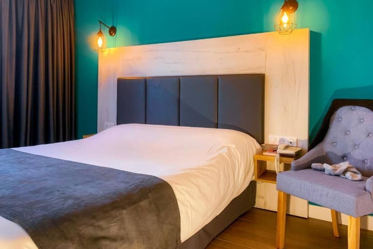 A hotel room with a bed, a chair, a nightstand, and a wall-mounted lamp.