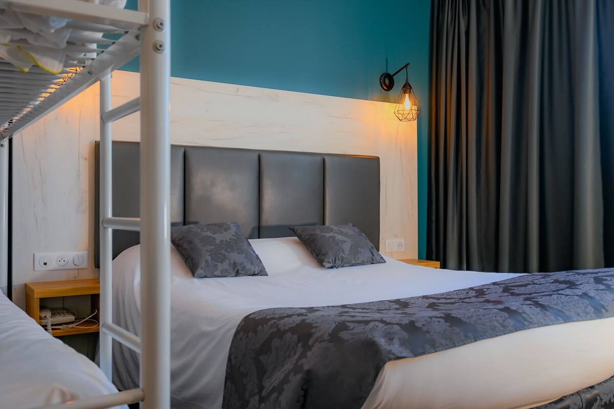 A hotel room with a bunk bed, a single bed, a nightstand with a phone, and a wall-mounted light fixture.