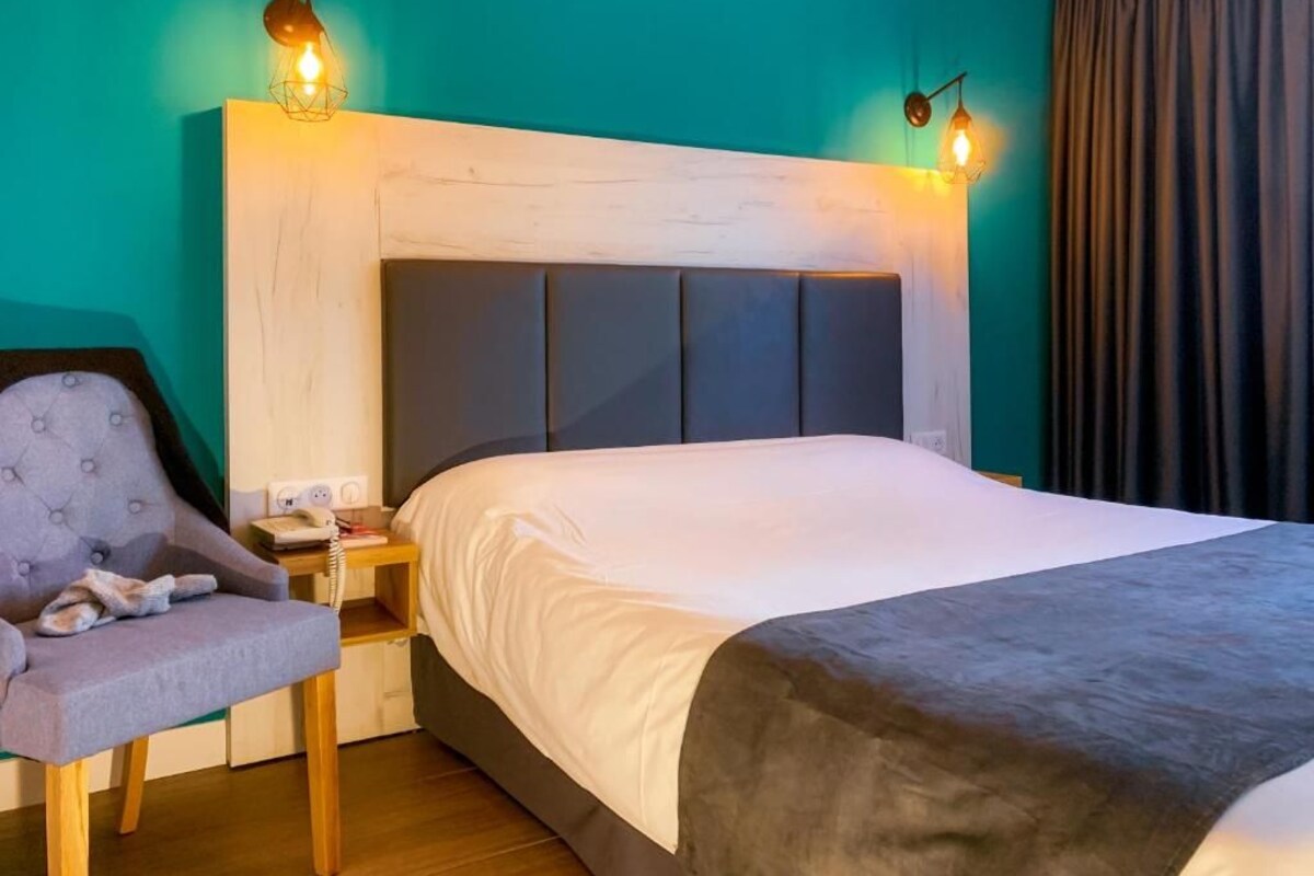A hotel room with a bed, a chair, a nightstand, and a wall-mounted lamp.