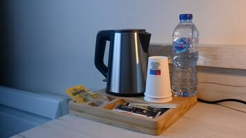 Een roestvrijstalen waterkoker, een witte beker, een waterfles en een dienblad met koffiepakketjes op een houten tafel.