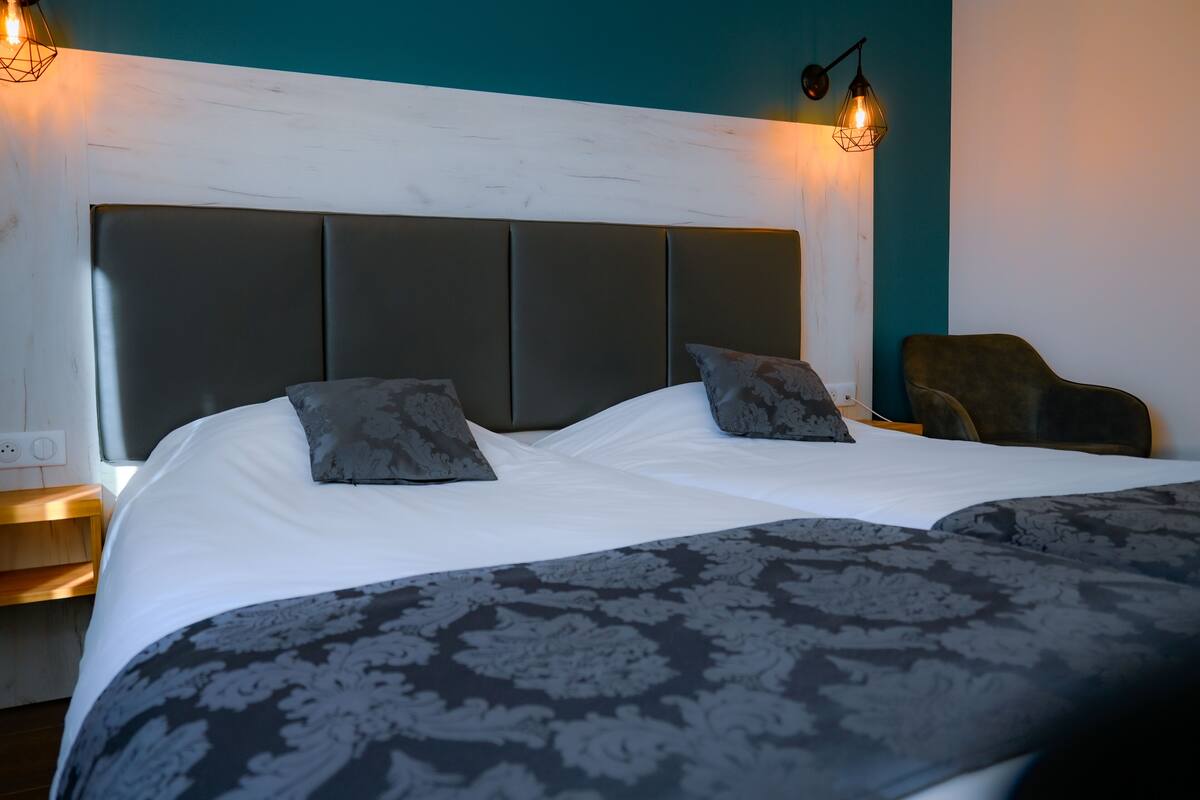 A double bed with a dark headboard, two pillows, a patterned bedspread, a nightstand with a lamp, and a chair.