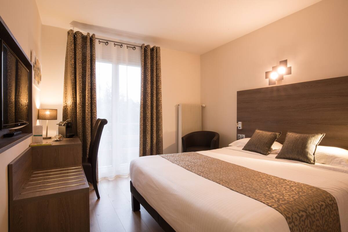 Habitación de hotel con una cama grande, un escritorio, una silla, un televisor y una ventana con cortinas.