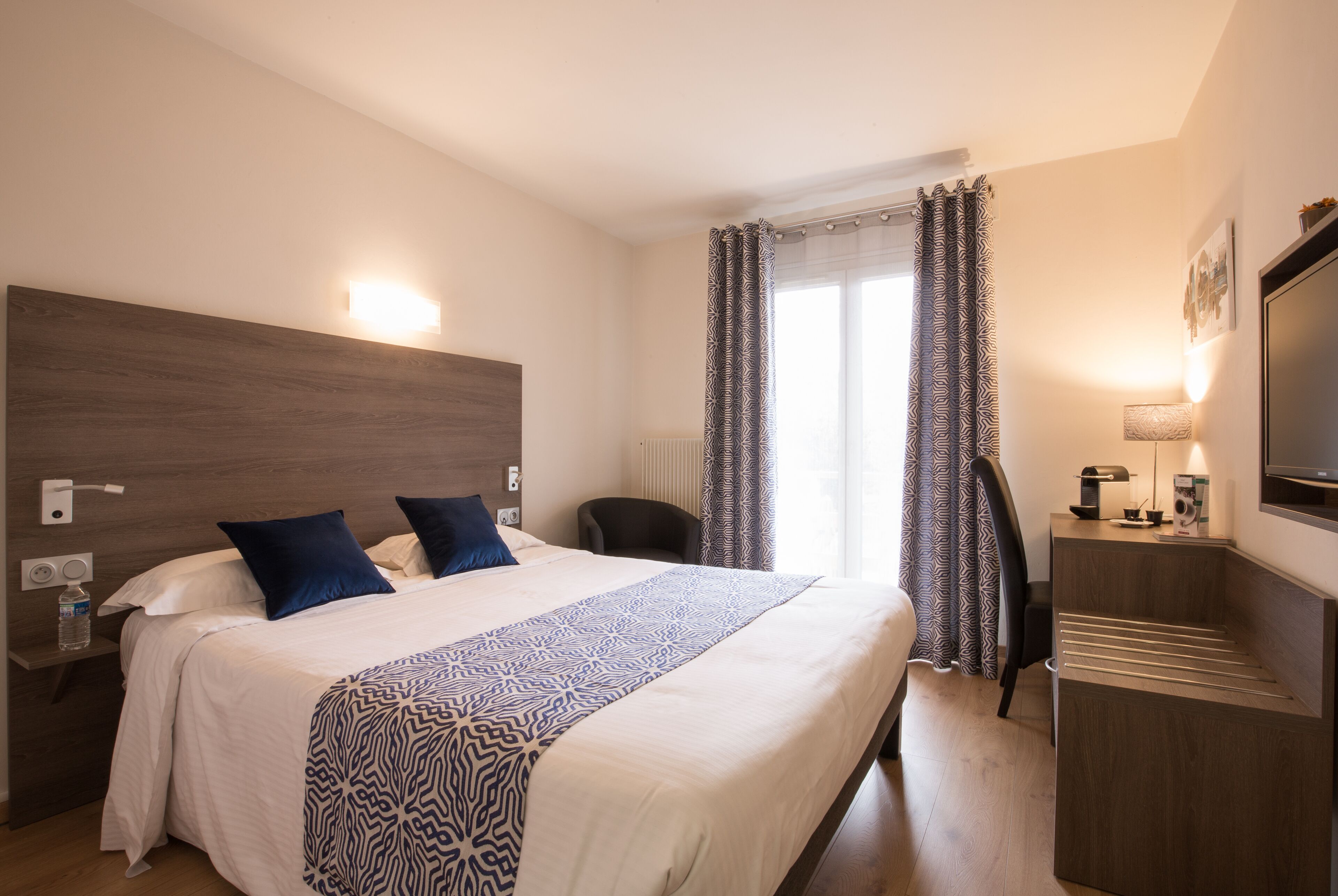 Une chambre d’hôtel avec un grand lit, un bureau, une chaise, une télévision et une fenêtre avec des rideaux.