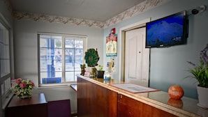 A reception area with a wooden desk, a mounted television, a window with blinds, and a decorative sign on the wall. - Travel Inn (Redding)