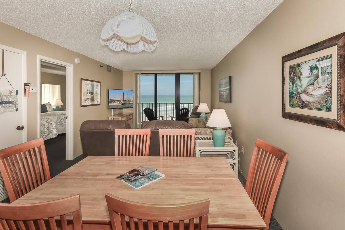 A dining area with a wooden table and chairs, a television, and a view of the ocean through large windows.