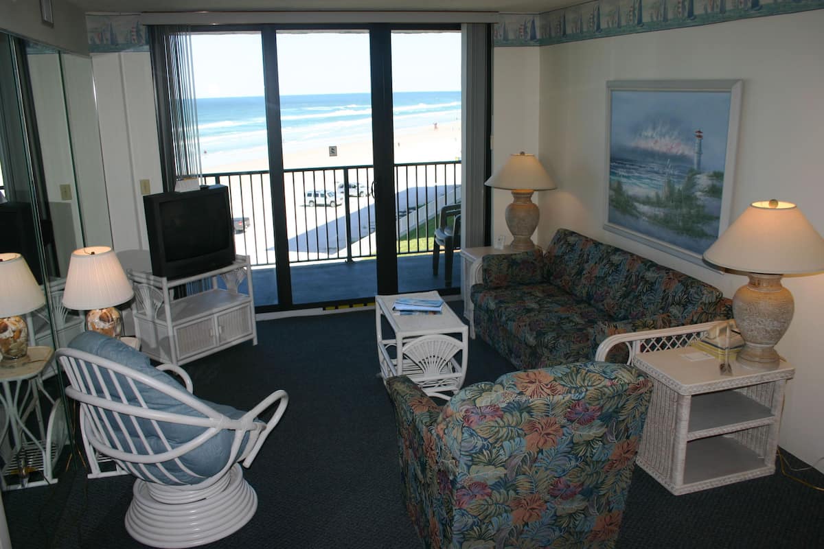 A room with a TV, two chairs, a sofa, and a table with lamps. A painting of a lighthouse is on the wall.