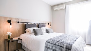 Ein modernes Schlafzimmer mit einem hölzernen Kopfteil, einem Nachttisch mit Lampe, einem Bett mit grauer Bettwäsche und einem Fenster mit weißen Vorhängen.